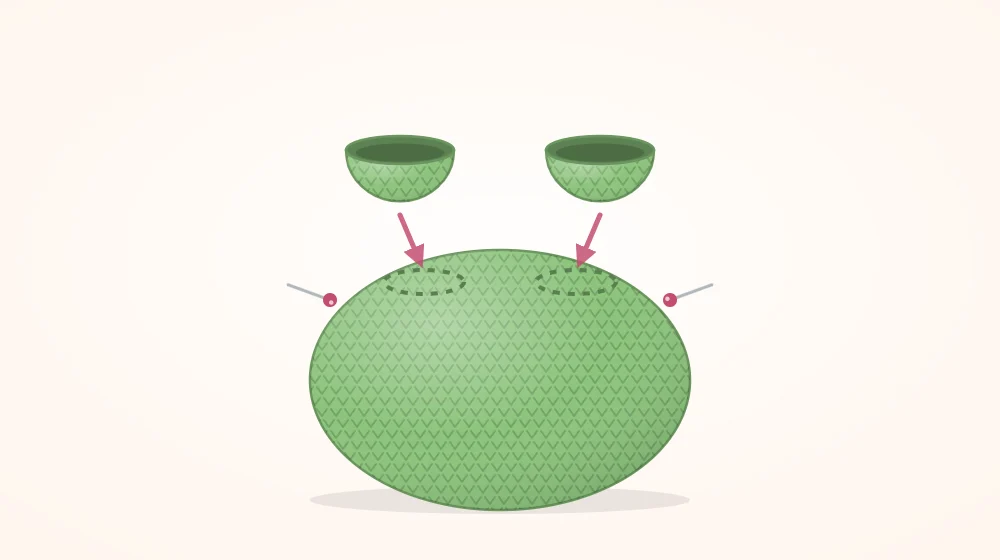Round green crocheted frog body with two small eye-bump domes held above it by pins, showing where they attach on top